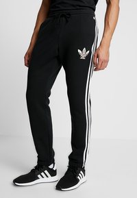 Man i svarta Adidas träningbyxor med vita ränder och logotyp, i kombination med svarta och vita Adidas sneakers, stående på grått golv.