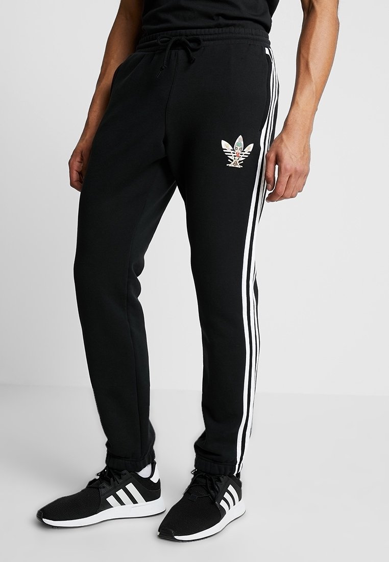 Man i svarta Adidas träningbyxor med vita ränder och logotyp, i kombination med svarta och vita Adidas sneakers, stående på grått golv.