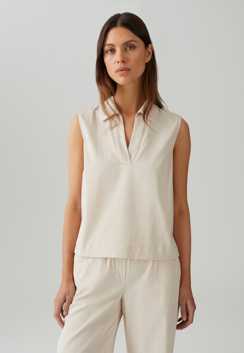 Mouwloze blouse in lichtbeige met een V-hals en subtiele textuur. Het ontwerp is ontspannen met een rechte zoom. Gecombineerd met bijpassende broeken.