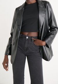 Svart kappa i konstläder med en strukturerad design, en enda knappstängning och två framfickor, kombinerad med högmidjade svarta jeans.