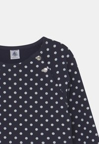 Mörkblå topp med vita prickar, rundad halsringning och två silverfärgade tryckknappar på axeln. Mjuk, strukturerad tyg.