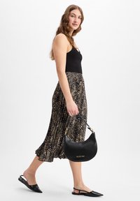 Zwarte lederen hobo tas met een gouden logo, gladde textuur en gebogen vorm. Het model draagt een zwarte top, een geplooide dierenprint rok en zwarte flats.