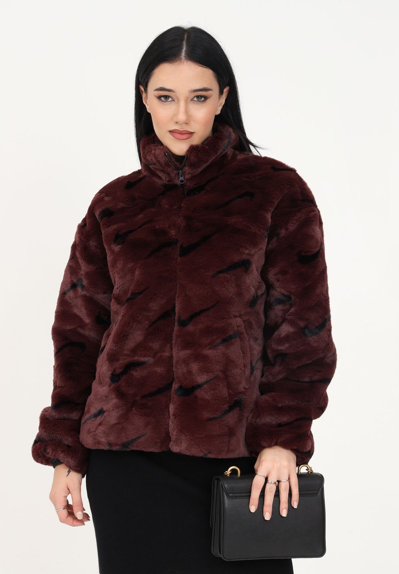 Veste en fausse fourrure bourgogne avec un col haut, présentant des motifs abstraits noirs, fermeture éclair et texture douce, assortie d'un sac à main noir.