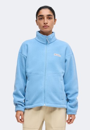 Ung person med mørkt hår i en blå fleecejakke med lynlås og lyse beige bukser, stående mod en ensfarvet lys baggrund.