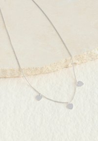 Strieborný náhrdelník s tromi malými príveskami v tvare srdca na jemnom retiazke, s lešteným povrchom. Pozadie je svetlé a textúrované.