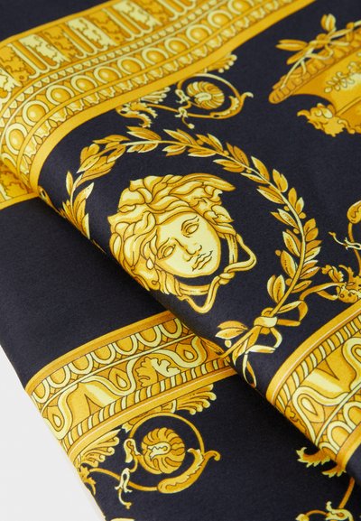 Černá tkanina se žlutými složitými vzory, která obsahuje klasické motivy, včetně obličeje a vavřínových věnců, přeložená přes sebe.