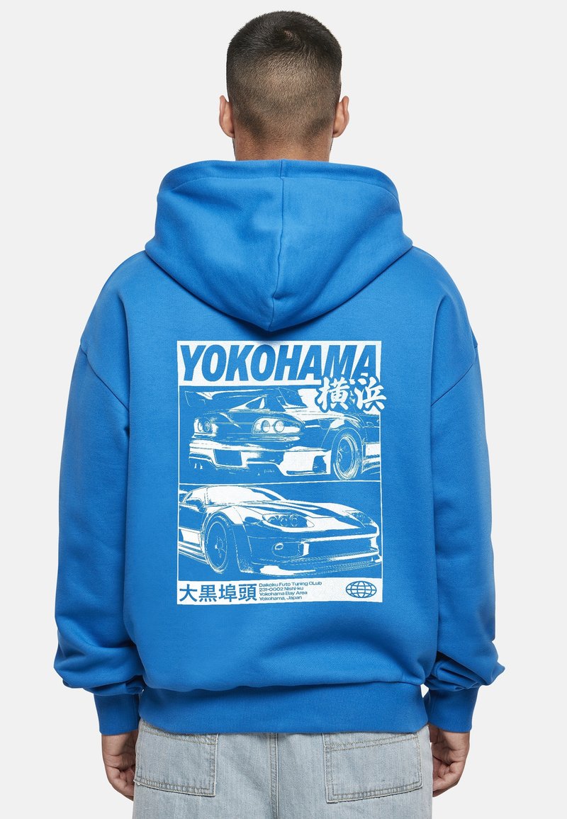 Modra jopa z kapuco z grafičnim potiskom avtomobilov. Material je mešanica bombaža. Na majici so besede "YOKOHAMA" in dodatni detajli v belem.
