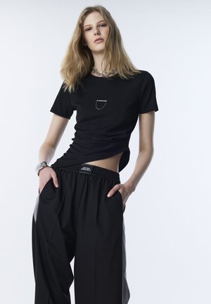 Giovane donna con lunghi capelli biondi che indossa una maglietta corta nera e pantaloni neri larghi con le mani nelle tasche, in posa su uno sfondo bianco.