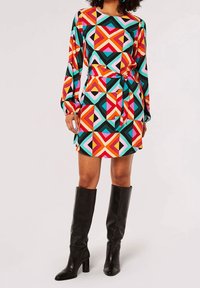 Buntes langärmliges Kleid mit einem geometrischen Muster in Rot, Pink, Türkis und Schwarz, mit gebundener Taille und ausgestellten Ärmeln. Schwarze kniehohe Stiefel.