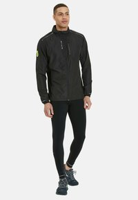 Black lightweight jacket with a high collar, zipper, and reflective accents, paired with black leggings and dark athletic shoes.