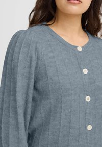 Šviesiai mėlynas kardiganas, pagamintas iš tekstūruoto audinio, su vertikaliais briaunelėmis, V formos iškirpte ir penkiomis kreminėmis mygtukais priekyje.