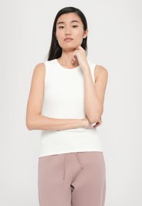 White sleeveless top with a ribbed texture, round neckline, and fitted design, paired with light pink sweatpants.
