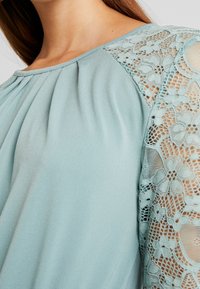 Blouse en bleu sarcelle clair avec un devant plissé et des accents en dentelle sur les manches. Le tissu semble doux avec une texture lisse.
