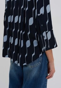 Navy blouse met een lichtblauw abstract patroon, lange mouwen, knoopmanchetten en een relaxte pasvorm, gecombineerd met lichtblauwe denimjeans.