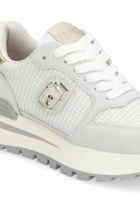 Zapatilla blanca con materiales de malla y ante, que presenta un logo metálico brillante, cordones blancos planos y una suela gruesa de color gris claro con tracción.