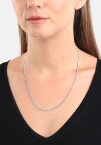 Donna che indossa una sottile collana d'argento sopra un top nero a V, con i capelli castani lisci visibili dietro le spalle.
