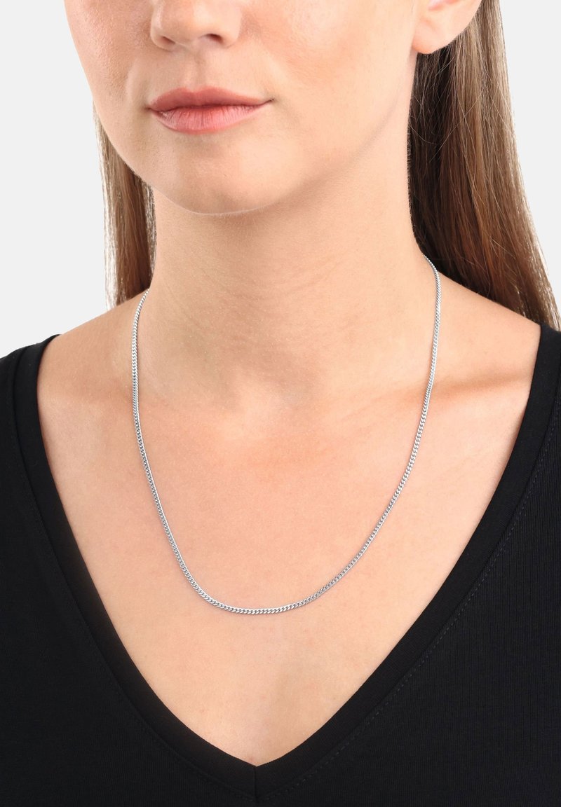 Donna che indossa una sottile collana d'argento sopra un top nero a V, con i capelli castani lisci visibili dietro le spalle.