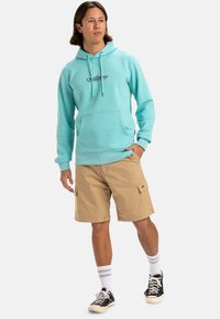 Sudadera azul clara con bolsillo frontal y cordones, combinada con pantalones cortos cargo beige, calcetines blancos a rayas y zapatillas negras.