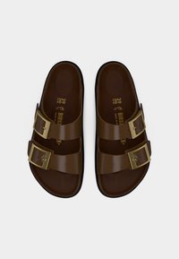 Καφέ σανδάλια Birkenstock με δύο ρυθμιζόμενες λουρίδες με αγκράφες και εσωτερικές ανατομικές σόλες, εμφανιζόμενα από πάνω σε λευκό φόντο.