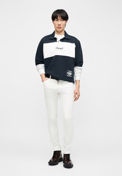 Jeune homme debout, portant un pull à manches longues noir et blanc, un pantalon blanc et des bottes marron foncé, sur un fond uni.