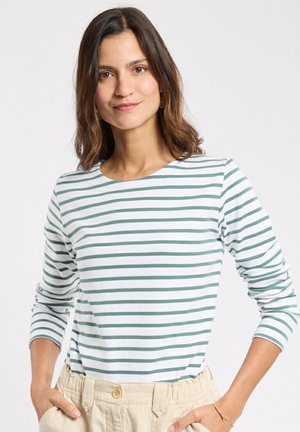 Femme portant une chemise blanche à manches longues avec des rayures horizontales vertes, un pantalon beige taille haute, les mains dans les poches, et un sourire subtil.