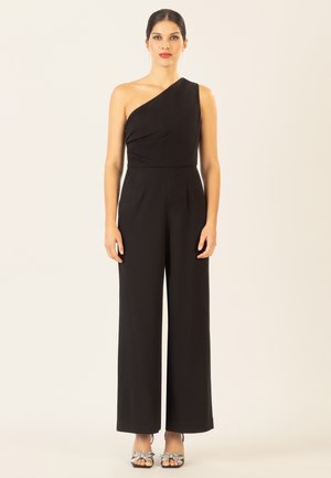 Woman wearing a black one-shoulder jumpsuit with wide legs and silver bow-detail high-heeled sandals, standing against a plain background.