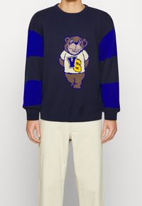 Pull en tricot bleu marine avec manches rayées bleues, arborant un motif d'ours ludique et "VS" sur une chemise blanche. Poignets et ourlet côtelés texturés.