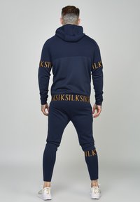 Tuta blu navy con dettagli in logo dorato, dotata di una felpa con cappuccio e pantaloni affusolati. Tessuto liscio, vestibilità elastica e polsini a coste. Calzature bianche.