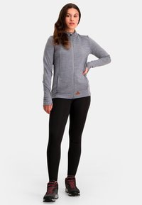 Grijze sportjack met ritssluiting en hoge kraag, gecombineerd met zwarte leggings en schoeisel met rode accenten. Soepel, lichtgewicht stof.