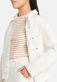 Hvit denimjakke med oransje sømmer, med krage, frontlommer og trykknapper, lagt over en krem- og tanstripet topp.