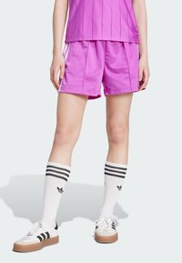 Roze atletske kratke hlače u kombinaciji s odgovarajućim topom, s glatkom teksturom i bočnim prugama. Nosene s bijelim čarapama do koljena i tenisicama.