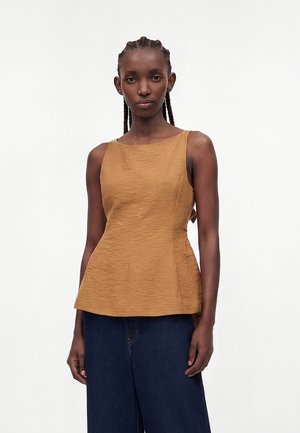 Jeune femme aux cheveux tressés portant un top sans manches texturé couleur moutarde et un pantalon large bleu foncé, debout devant un fond uni.