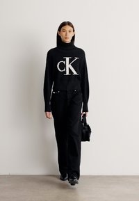 Maglione nero a collo alto in maglia con grafica bianca "ck", abbinato a pantaloni neri a vita alta. La modella tiene una piccola borsa nera.