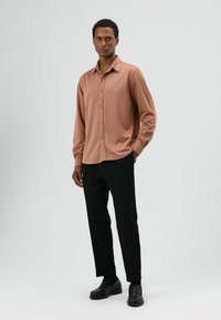 Langärmeliges, hellbraunes Hemd mit Knopfleiste, kombiniert mit schwarzen Hosen und schwarzen Loafers. Glatter Stoff, strukturierte Passform und klare Linien.