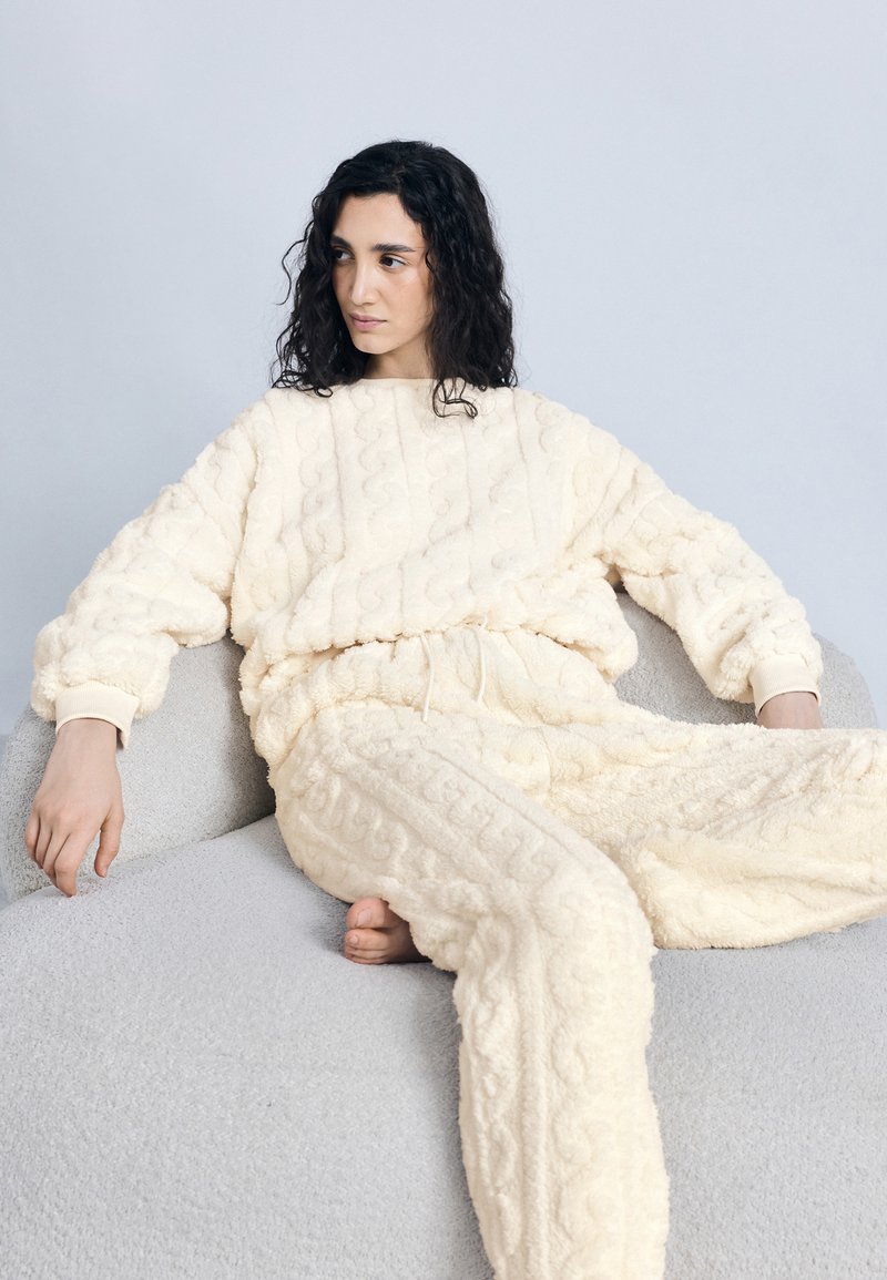 Γυναίκα με σκούρα, σγουρά μαλλιά, ντυμένη με κρεμ υφασμάτινα ρούχα για χαλάρωση, κάθεται ξυπόλυτη σε απαλό γκρι μαλακό κάθισμα, μπροστά σε ανοιχτόχρωμο τοίχο.