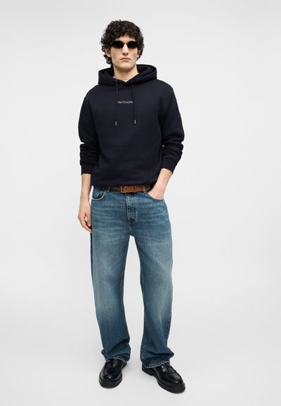 Jeune homme portant un sweat à capuche noir, un jean bleu, une ceinture marron, des chaussures noires et des lunettes de soleil, debout avec une main dans la poche sur un fond uni.