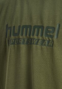 Oliwkowa koszulka z teksturowanym wykończeniem; na przodzie znajduje się ciemnozielony nadruk z napisem "hummel SPORTSWEAR".