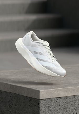 Chaussure de sport blanche avec une tige texturée, trois bandes latérales, une semelle amortie et un bout arrondi, posée au-dessus d'une surface en béton.