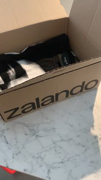 Dėžė iš kartono su "zalando" ženklinimu, iš dalies atidaryta, atidengiant viduje sulankstytus drabužius, ant šviesios marmurinės paviršiaus.