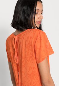 Blusa laranja transparente com um padrão ondulado, mangas curtas, decote redondo e um fecho de laço nas costas. Textura suave e material leve.