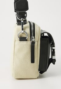Canvas bag in beige with black leather accents, featuring a zippered closure, side pocket, and adjustable strap with silver hardware.