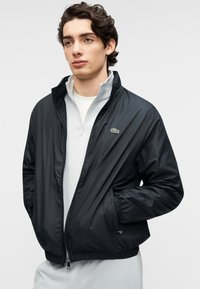 Μαύρο ελαφρύ μπουφάν με φερμουάρ μπροστά, ελαστικά μανίκια και τελείωμα. Διαθέτει μικρό πράσινο λογότυπο στο αριστερό στήθος, φορεμένο πάνω από ανοιχτόχρωμο τοπ.