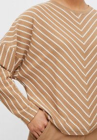 Pull en tricot marron avec des rayures en zigzag blanches, encolure ronde et coupe décontractée. Tissu texturé avec des manches longues et des détails aux poignets.
