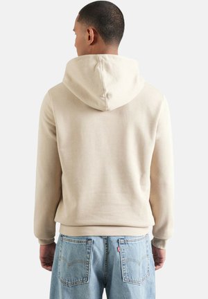 Sudadera con capucha de color beige y diseño ajustado, que incluye capucha, puños y dobladillo acanalados. Se muestra desde atrás, combinada con jeans vaqueros de mezclilla azul claro.