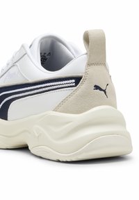 Puma sneaker med en hvid syntetisk overdel, marineblå accenter og beige suede hæl. Tykk, tekstureret sål med udtalt bølgedesign.