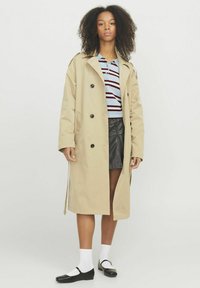 Cappotto beige con design doppiopetto, grandi bottoni neri e spalline. Indossato sopra una maglietta a righe e una gonna di pelle.