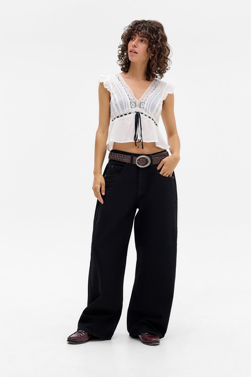 Mlada žena s kovrčavom kosom nosi bijeli crop top s čipkastim rubom, široke crne hlače, smeđi remen i tamne mokasine, stoji na bijeloj pozadini.