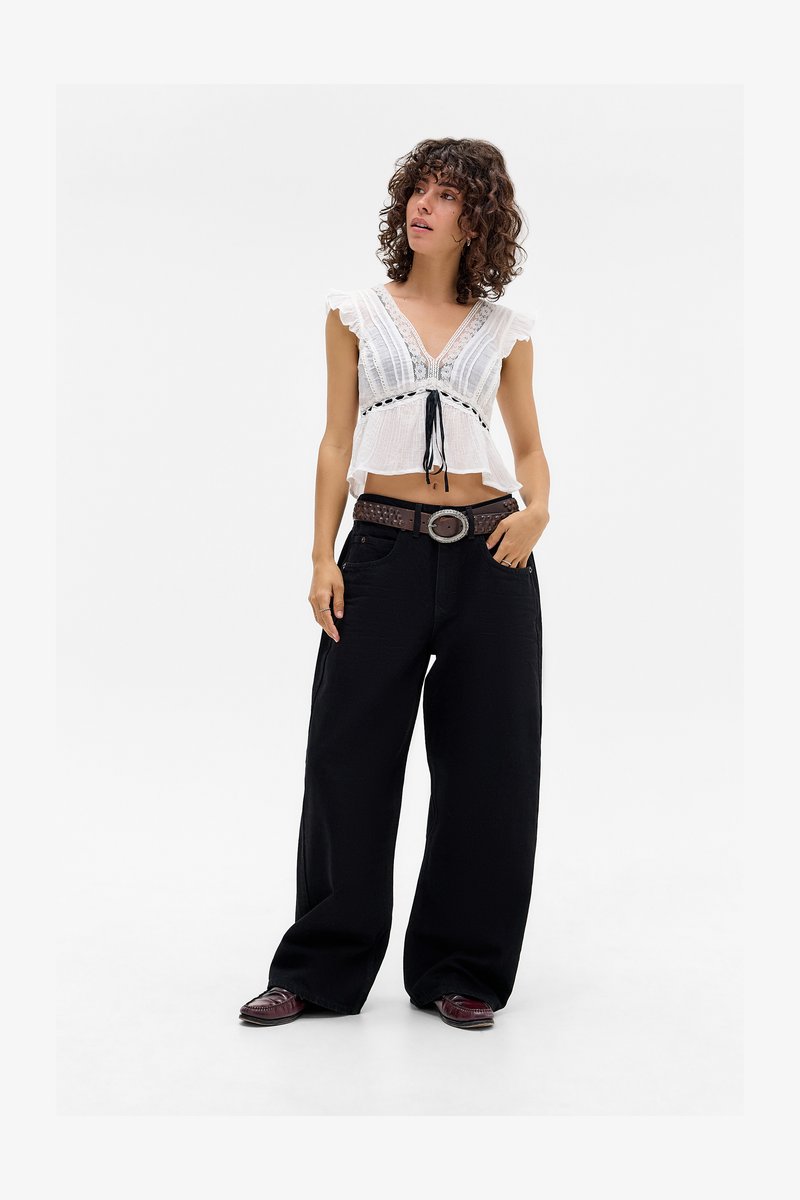 Mlada žena s kovrčavom kosom nosi bijeli crop top s čipkastim rubom, široke crne hlače, smeđi remen i tamne mokasine, stoji na bijeloj pozadini.