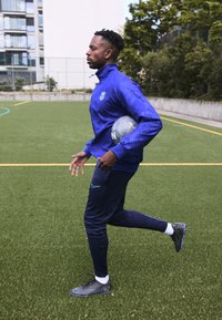 Chaqueta deportiva azul con logo, pantalones de chándal azul marino y botas de fútbol negras. Sosteniendo un balón de fútbol gris, de pie sobre césped artificial.