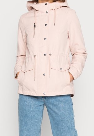 Mujer con chaqueta con capucha de color rosa claro con cierres de botones y cremallera y dos bolsillos delanteros con solapa, combinada con vaqueros azules.